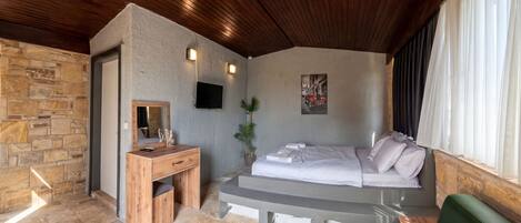 Quarto com parede de pedra, teto de madeira, uma cama, escrivaninha, espelho, planta em vaso e televisão.