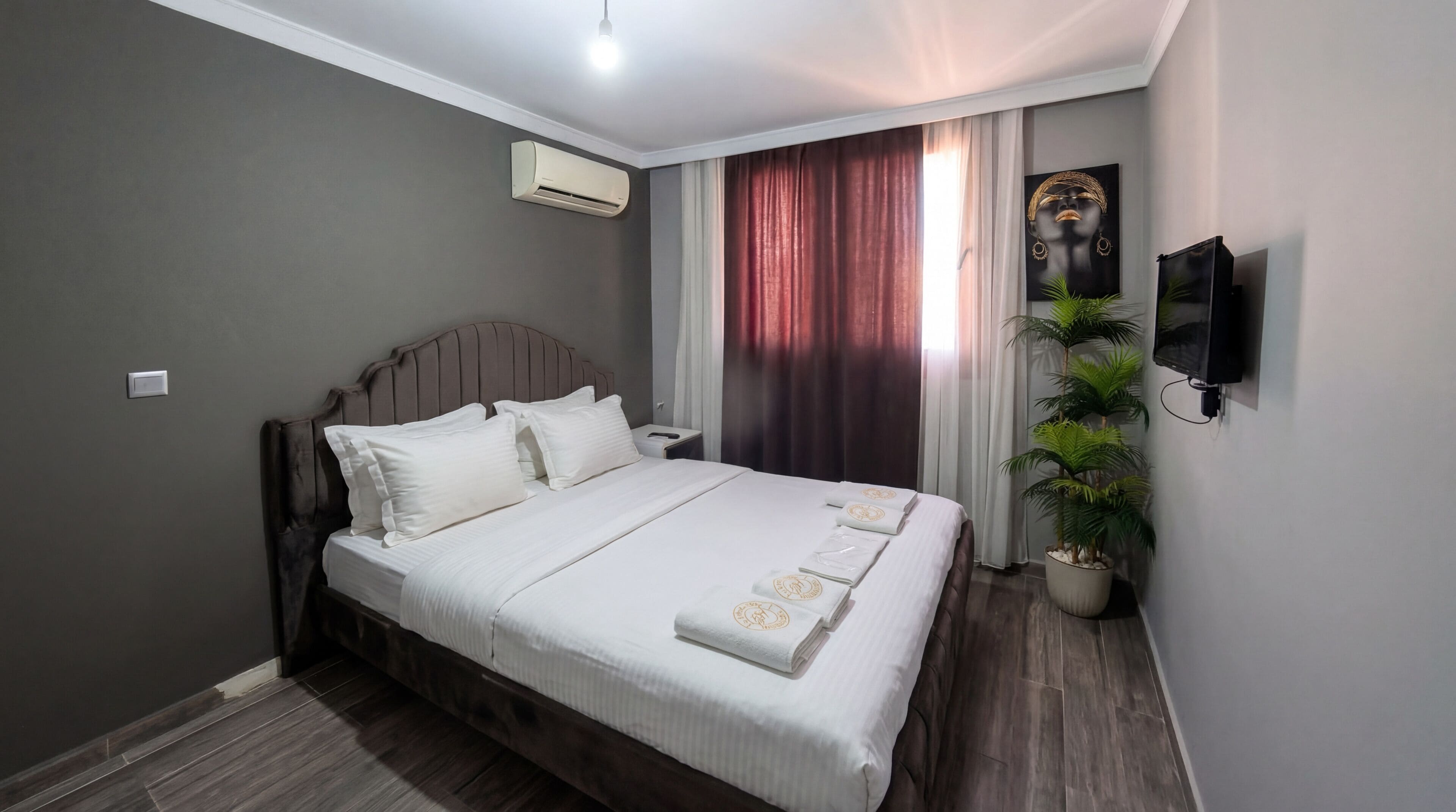 Una cama bien hecha con ropa blanca y una almohada decorativa, una mesita de noche con lámpara, un televisor montado en la pared y una planta en maceta.