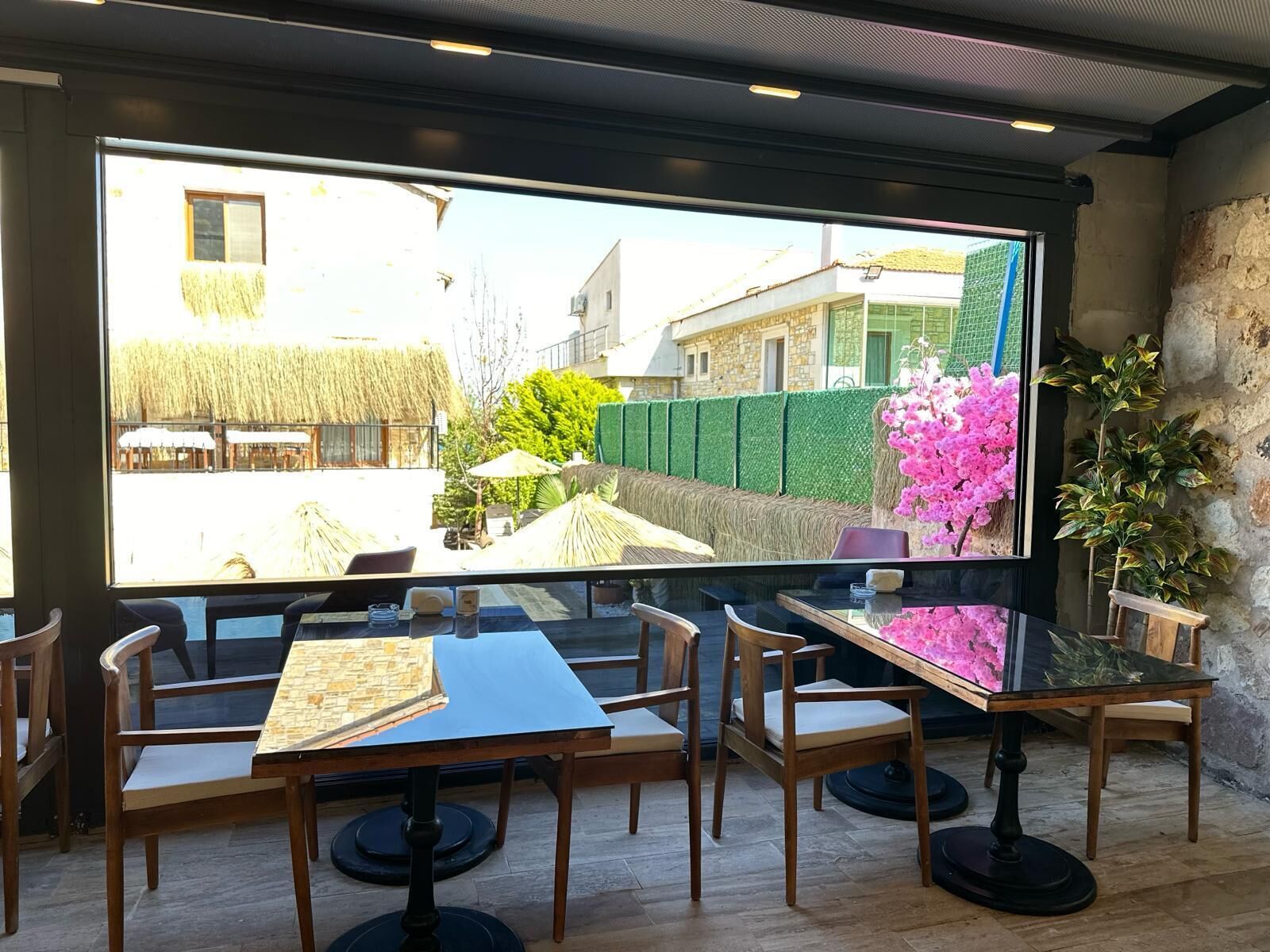 Un comedor con un ventanal grande que da a un jardín y edificios. Hay varias mesas y sillas de madera, y una planta en maceta es visible a la derecha.