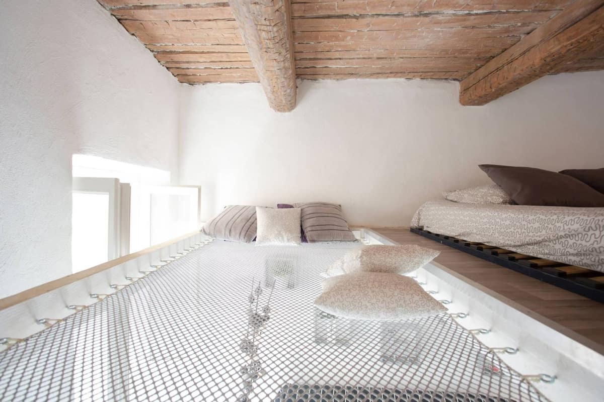 Une pièce avec un plafond haut, un lit recouvert d’une literie à motifs, un petit canapé et un sol en treillis blanc.
