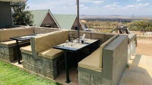 An outdoor patio with cushioned seating and a table, an umbrella, and a view of a vineyard and distant hills. - Outback Kenya Lodge (Machakos)