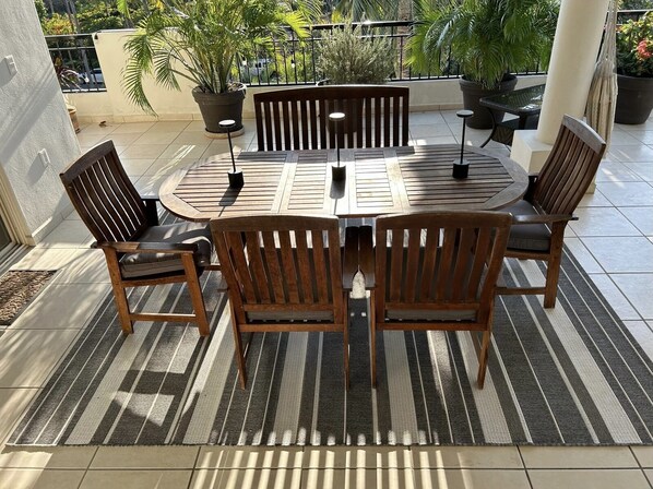 Une table ronde en bois, entourée de six chaises, de plantes en pot et d’un tapis à rayures, le tout sur un balcon.