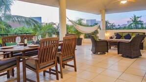 Eine Terrasse im Freien mit Holzmöbeln, einer Hängematte und Blick auf Palmen und Gebäude.