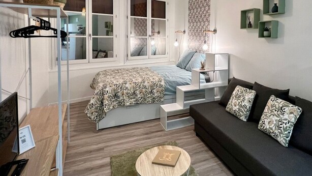 A compact bedroom with a bed, a sofa, a small table, and a window with curtains. - 🌿 L'Oliveraie 🌿 (Brioude)