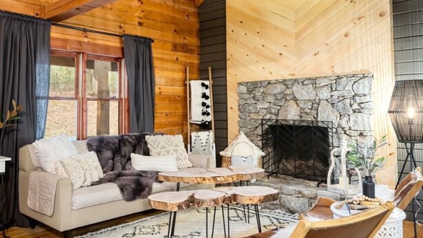 A cozy cabin interior with a stone fireplace, wooden beams, a sofa, and a dining table. - Luxury Stay: HGTV Log Cabin*Hot tub*Arcade*EV*Fire Pit*Nature (Fairview)