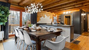 A wooden interior with a dining table set for six, a kitchen area, and a large window overlooking a forest. - Luxury Stay: HGTV Log Cabin*Hot tub*Arcade*EV*Fire Pit*Nature (Fairview)