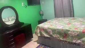Une chambre avec des murs verts, un lit recouvert d’une couvre-lit à motifs floraux, une commode avec un miroir, une lampe et un rideau.