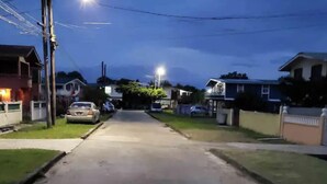 Une rue résidentielle au crépuscule, bordée de maisons des deux côtés, avec des lampadaires et des voitures garées.