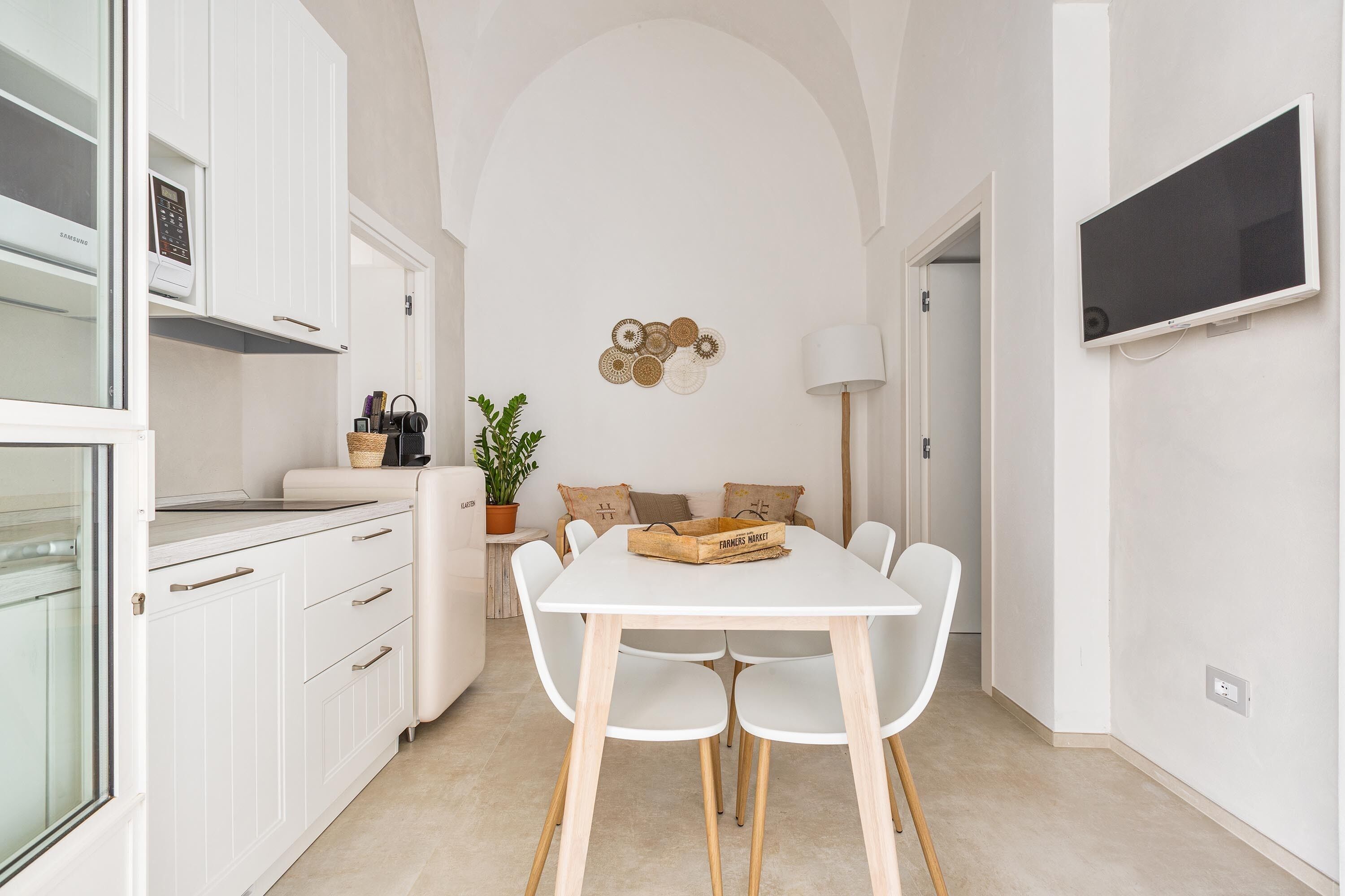 A compact kitchen with white cabinetry, a dining table with four chairs, a potted plant, and a wall-mounted TV.