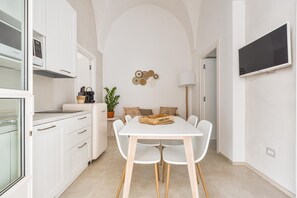 Cozinha compacta com armários brancos, uma mesa de jantar com quatro cadeiras, uma planta em vaso e uma TV fixada na parede.