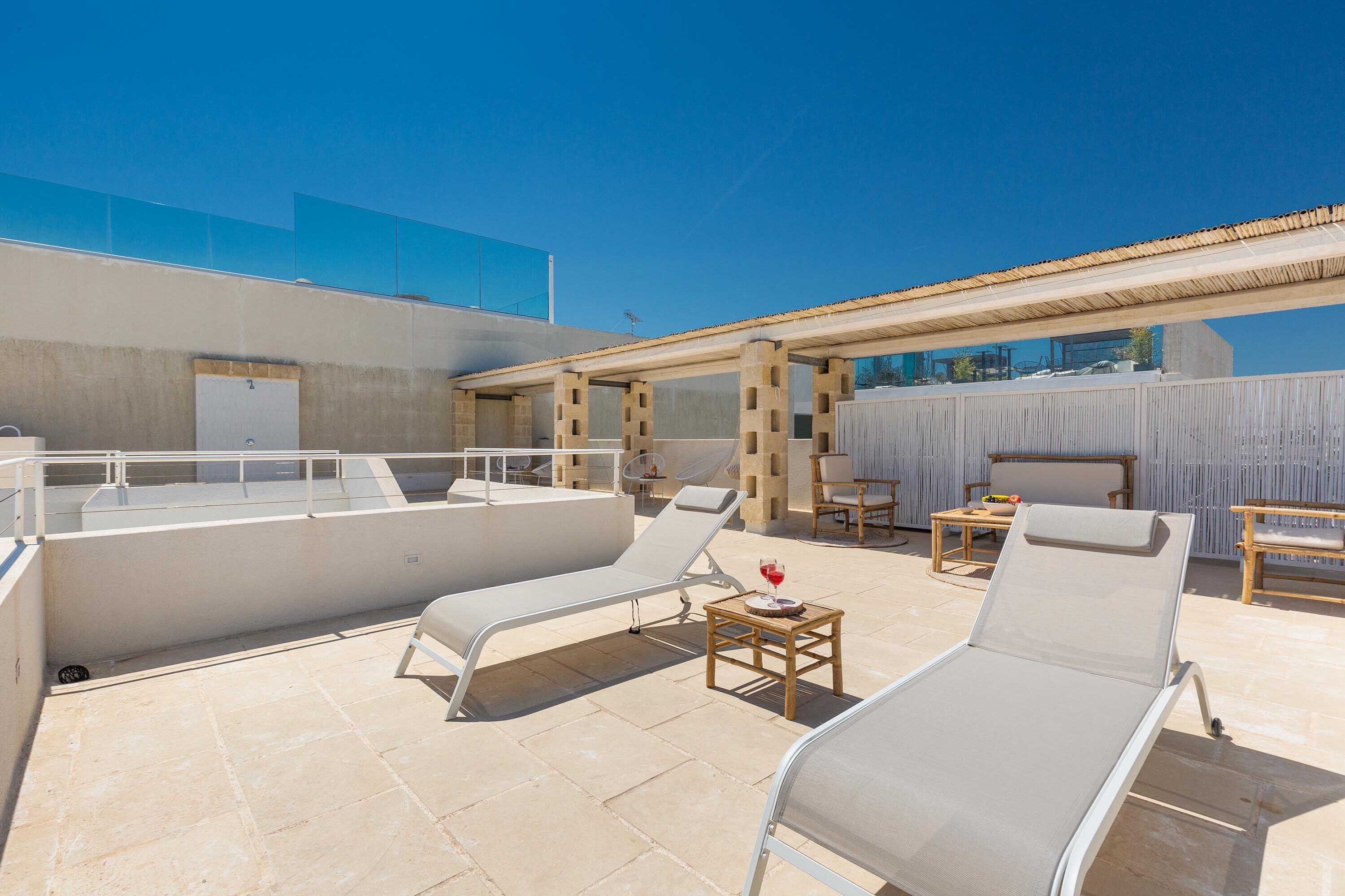 A rooftop terrace with lounge chairs, a table with a glass of red wine, and a covered seating area.