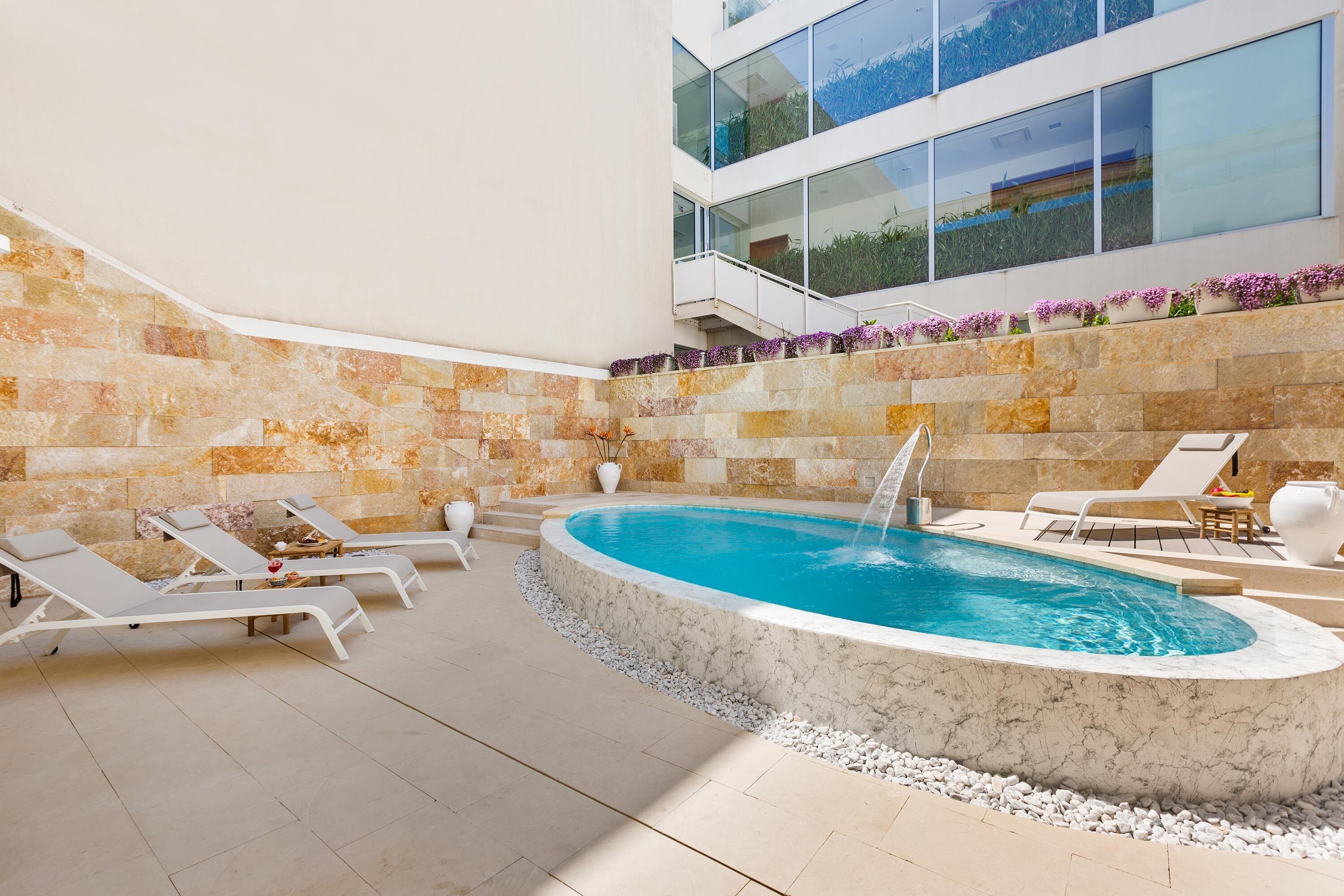 A modern indoor pool area with lounge chairs, a hot tub, and a stone wall.