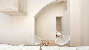 Un área de estar moderna al aire libre con sofás y sillones blancos acolchados, una peque ña mesa con bebidas y pasteles, y una unidad de aire acondicionado grande montada en la pared.