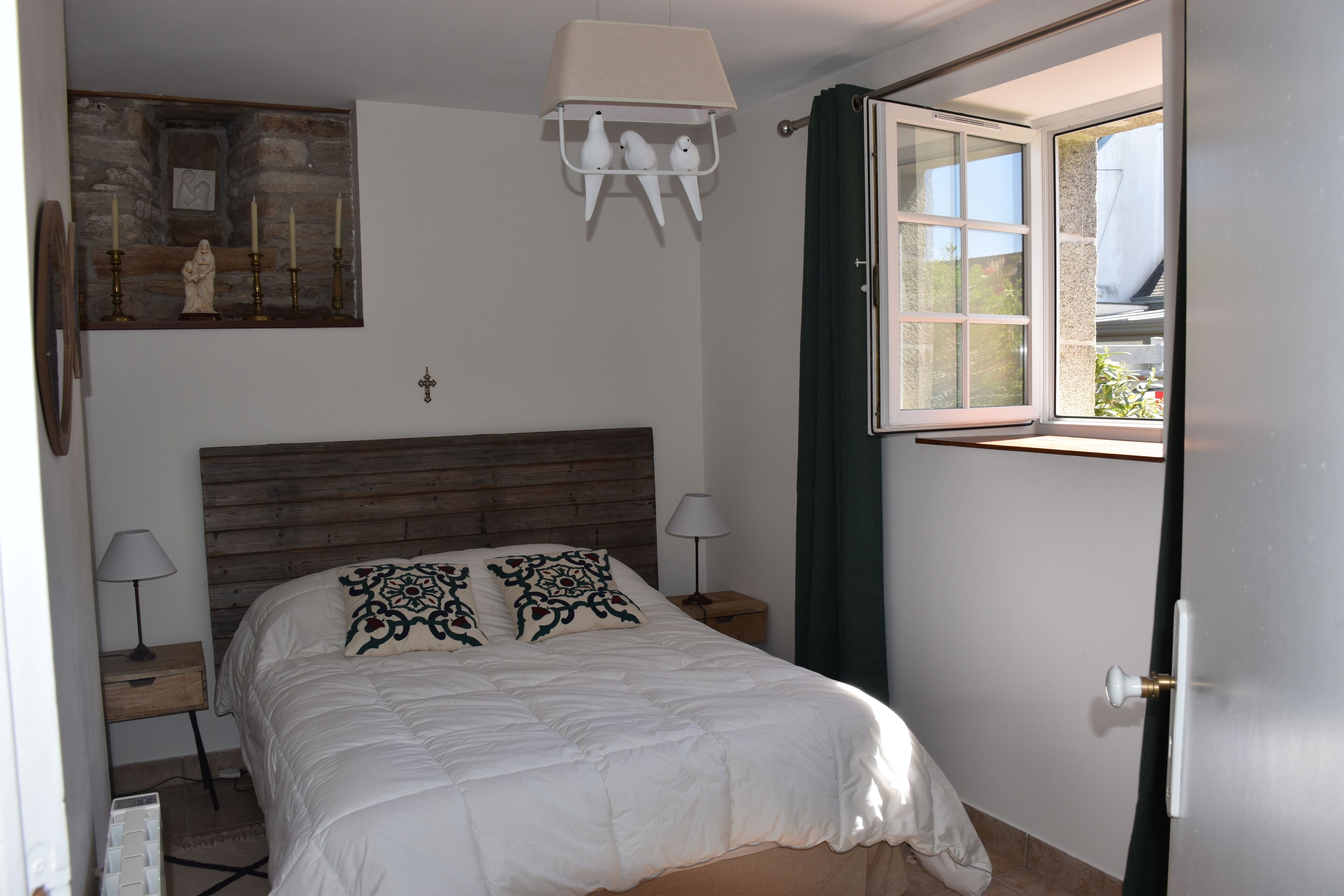 A bedroom with a wooden headboard, a bed with white bedding, two bedside lamps, a window with curtains, and a stone wall with a religious statue.