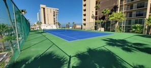 A tennis court with a net, surrounded by a green fence, palm trees, and a building in the background. - Spi Fun And Sun 2 Bedroom Condo by RedAwning (South Padre Island)