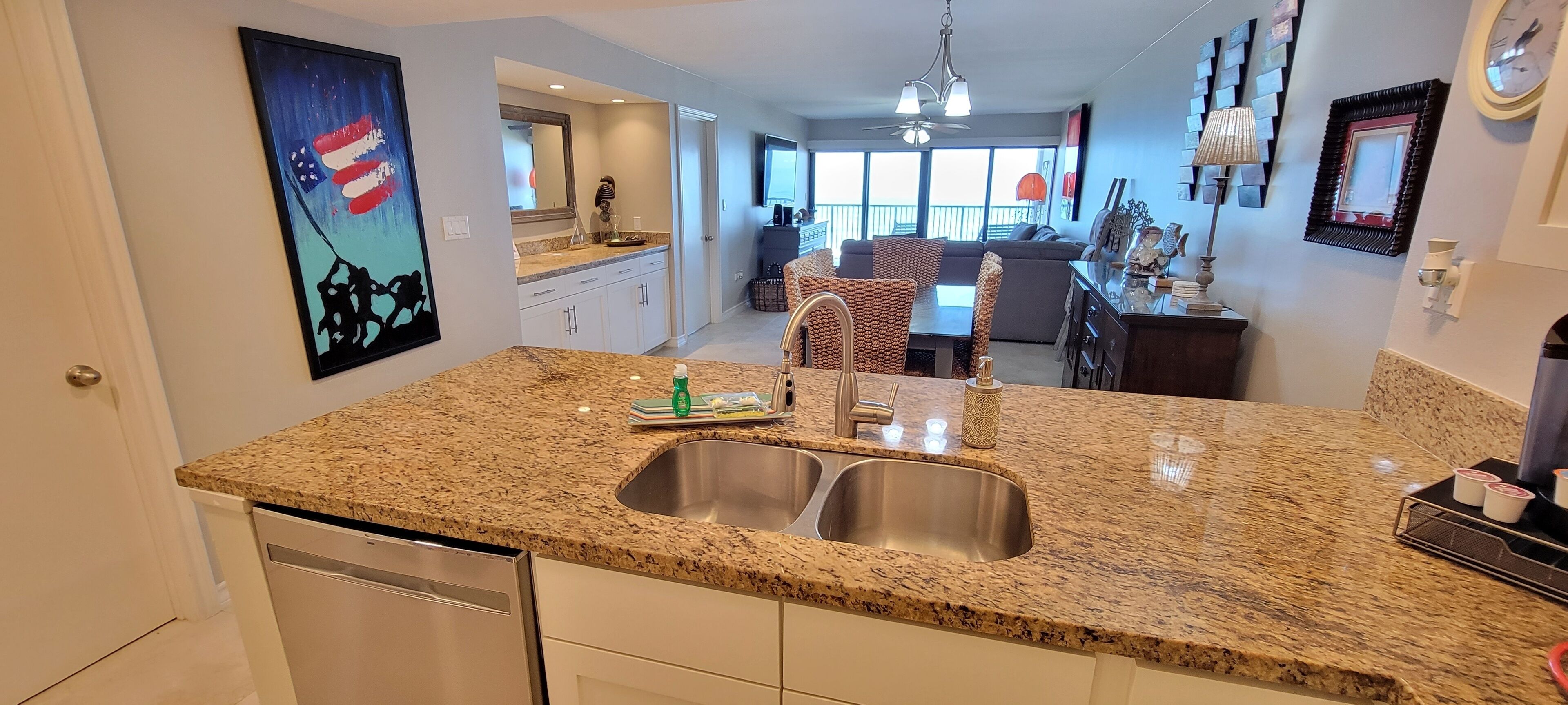 A kitchen with granite countertops, stainless steel sinks, and white cabinetry. There is a dining area with wicker chairs and a chandelier.