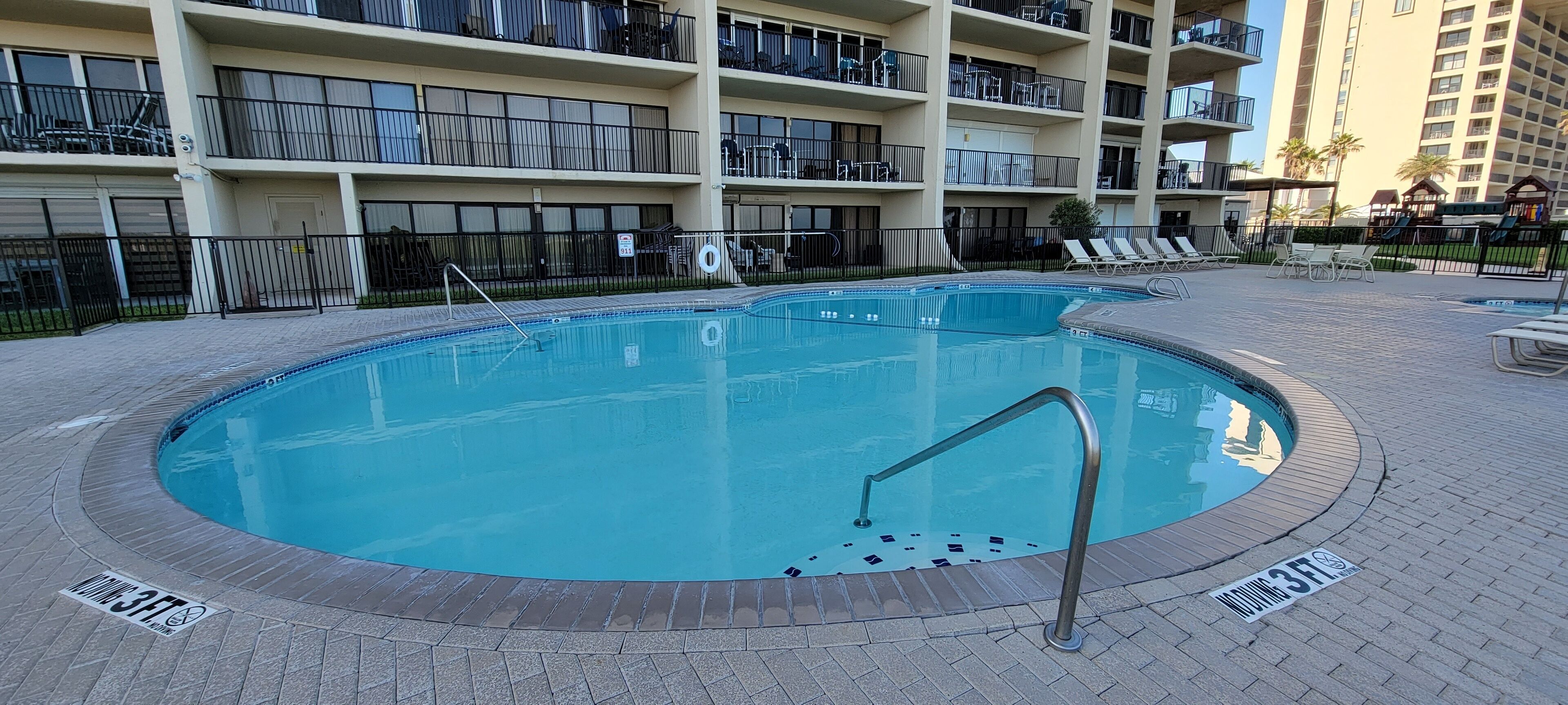 A swimming pool with a metal handrail, surrounded by a brick deck and a fence.