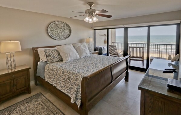 A bedroom with a large bed, a ceiling fan, a nightstand, a desk, and a view of the beach through large windows. - Spi Fun And Sun 2 Bedroom Condo by RedAwning (South Padre Island)