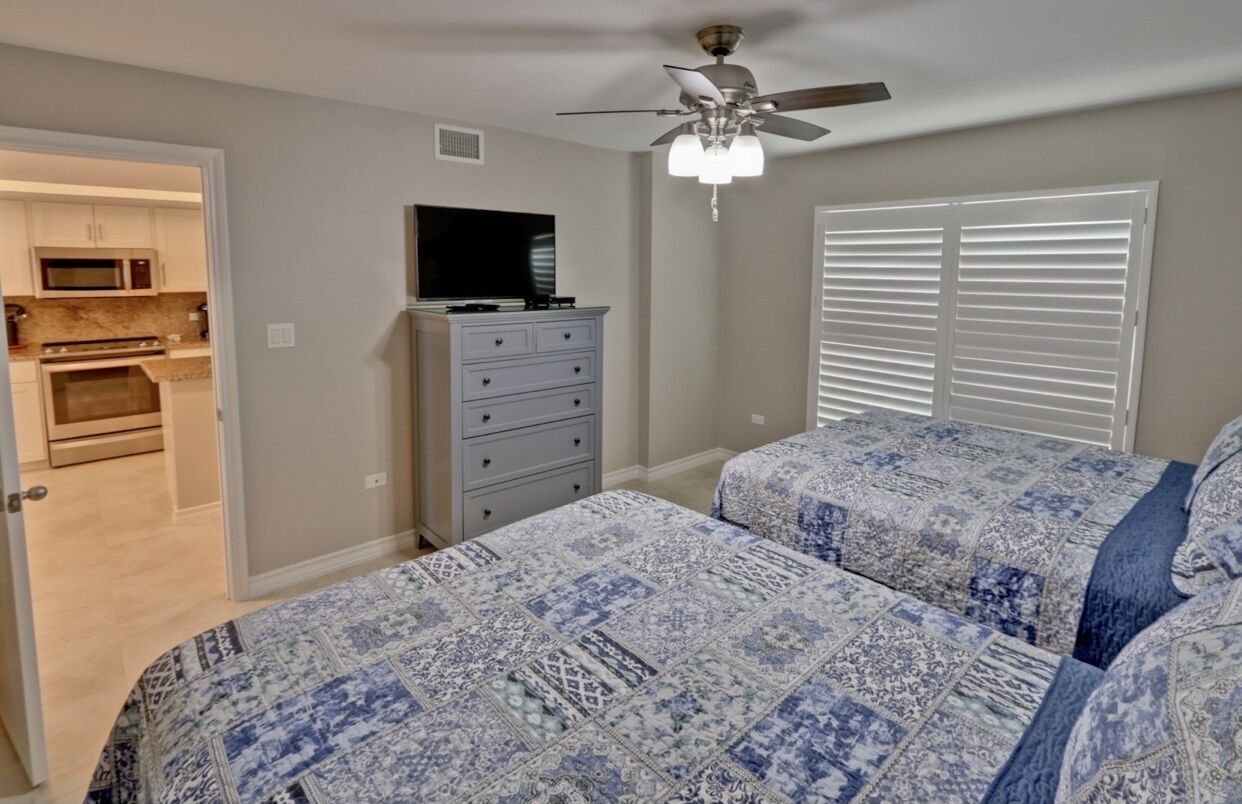 A bedroom with two beds, a ceiling fan, a TV on a dresser, and a view of the kitchen through an open door.