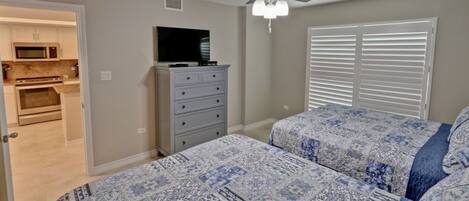A bedroom with two beds, a ceiling fan, a TV on a dresser, and a view of the kitchen through an open door.