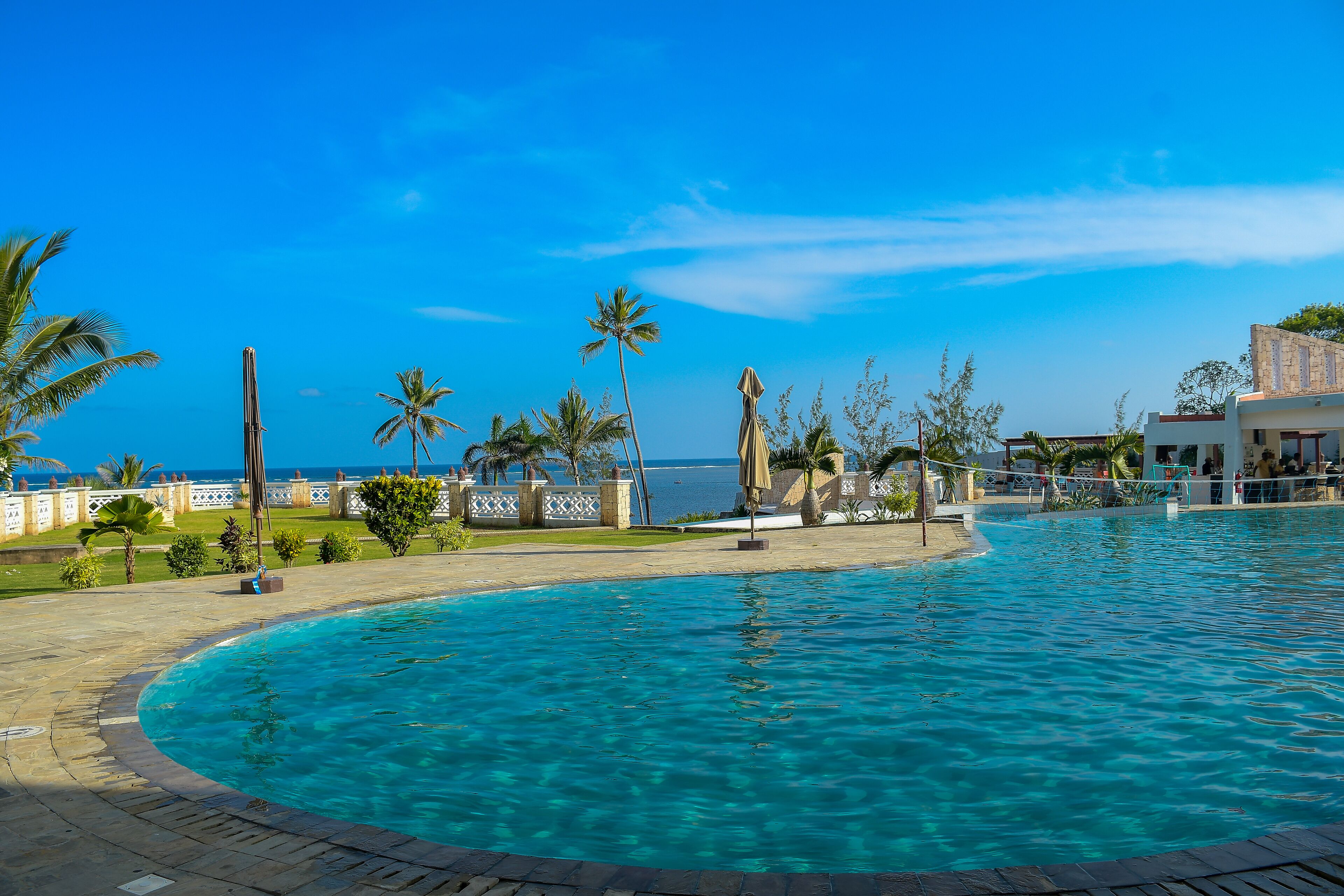 Piscina com vista clara do oceano, cercada por palmeiras e um edifício com fachada branca.