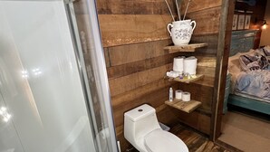 A bathroom with a glass-enclosed shower, a toilet, and a sink. There is a wooden wall paneling and a shelf with toiletries. - COZY RIVER FRONT CABIN (Blue Ridge)