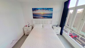 A bedroom with a large bed, two bedside tables, a nightstand, a window with a view of a red car, and a sunset painting on the wall.
