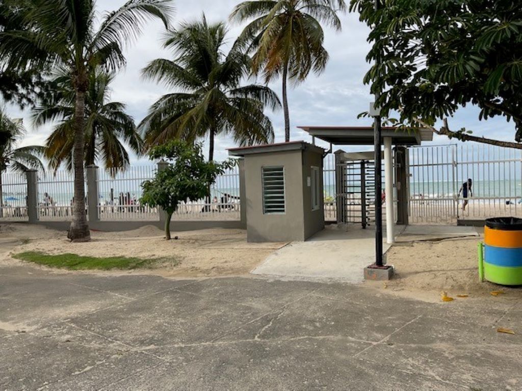 Une zone située au bord de la plage, avec un chemin en béton, des palmiers et une entrée fermée par un portail.