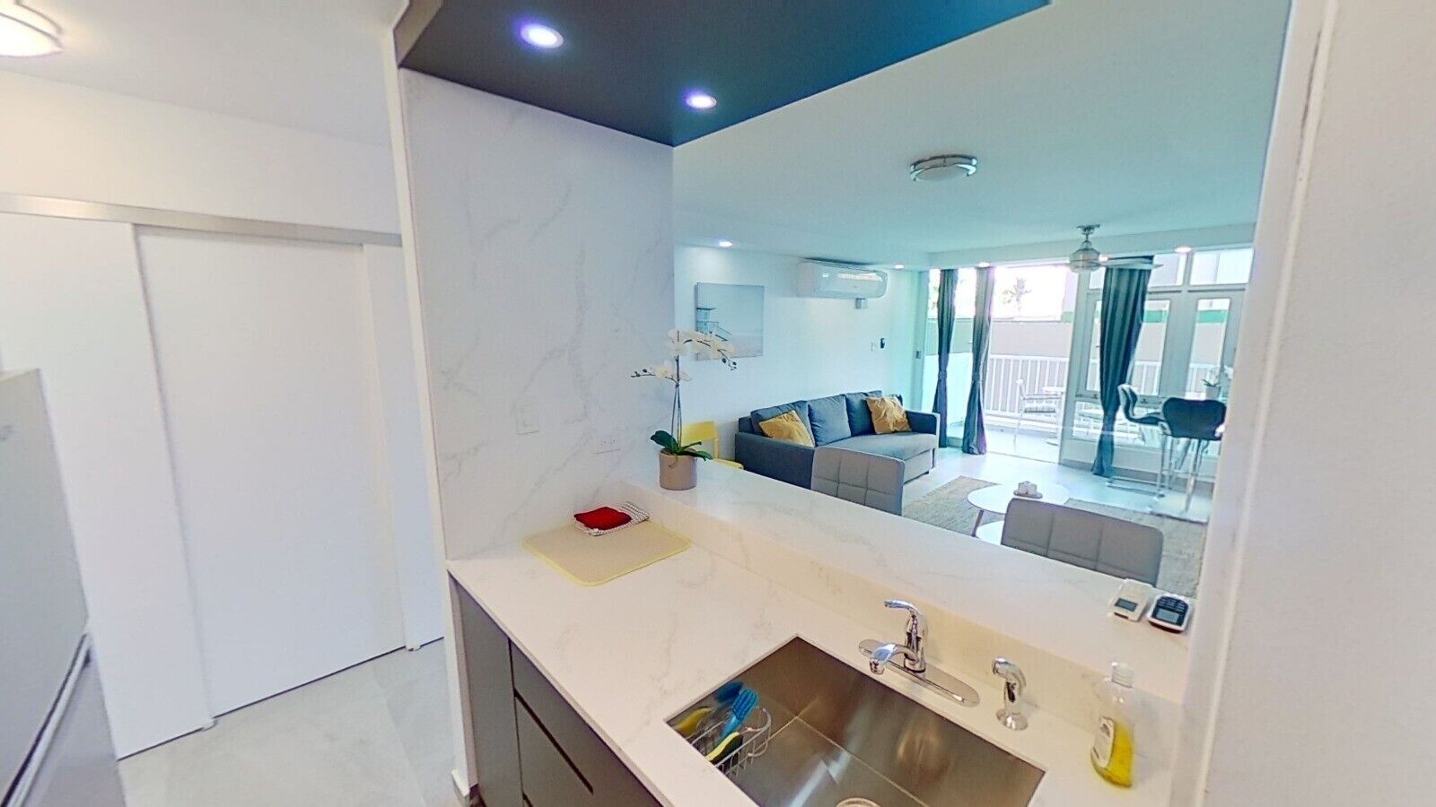Cocina moderna con encimera blanca, pileta de acero inoxidable y un espejo grande que refleja una zona de estar con sofá y mesa de comedor.
