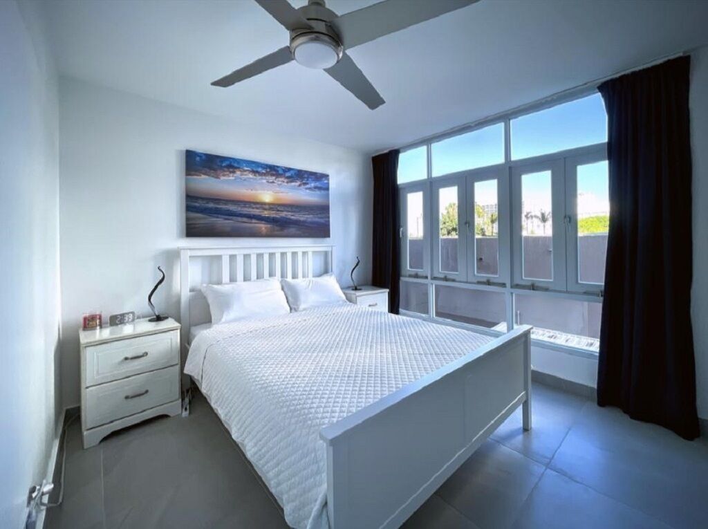 Une chambre à coucher comprenant un lit, une table de chevet, un ventilateur de plafond, une fenêtre avec des rideaux et un tableau accroché au mur.