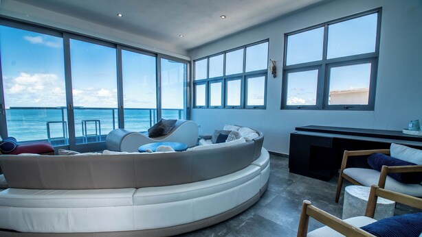 Un salón moderno con un sofá curvo, una silla de madera y un escritorio. Amplias ventanas ofrecen vistas al océano.