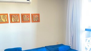 Un salon moderne avec un canapé d’angle bleu, un climatiseur au plafond et une fenêtre ornée de rideaux blancs.