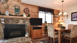 A cozy living room with a stone fireplace, wooden walls, a television, a dining table with patterned chairs, and a chandelier. - Seven Springs 3 Bedrooms Premium Condo by RedAwning (Champion)