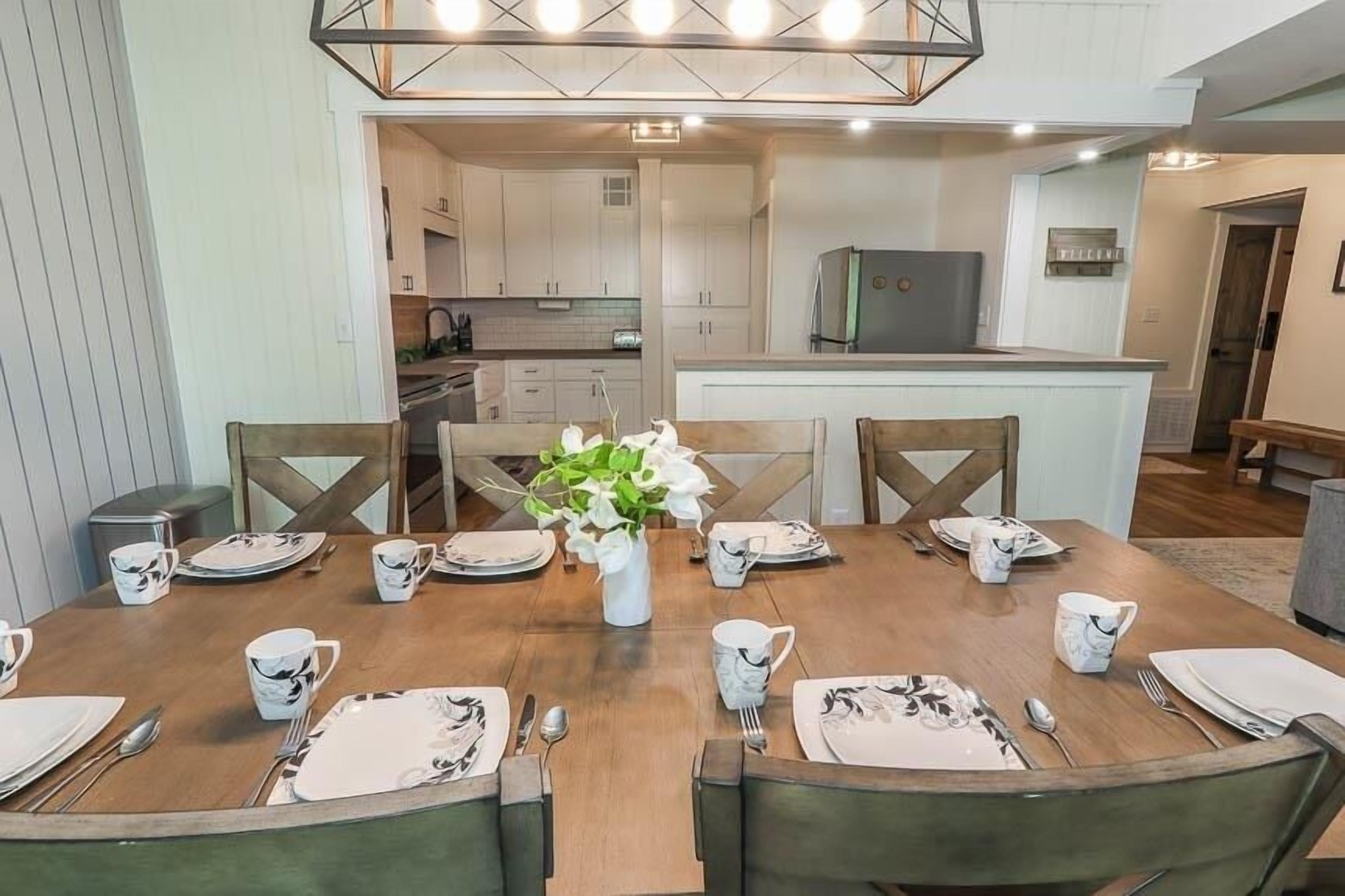 A dining area with a wooden table set for four, surrounded by chairs. A vase with white flowers is placed in the center.