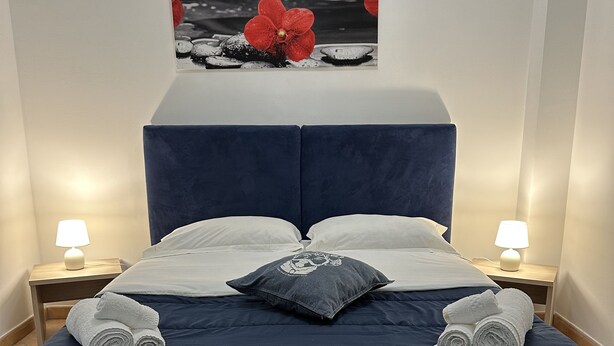 A neatly made bed with a blue headboard, white pillows, and rolled towels. There are two bedside tables with lamps, and a framed picture above the bed. - CARTARI APARTMENT (Palermo)