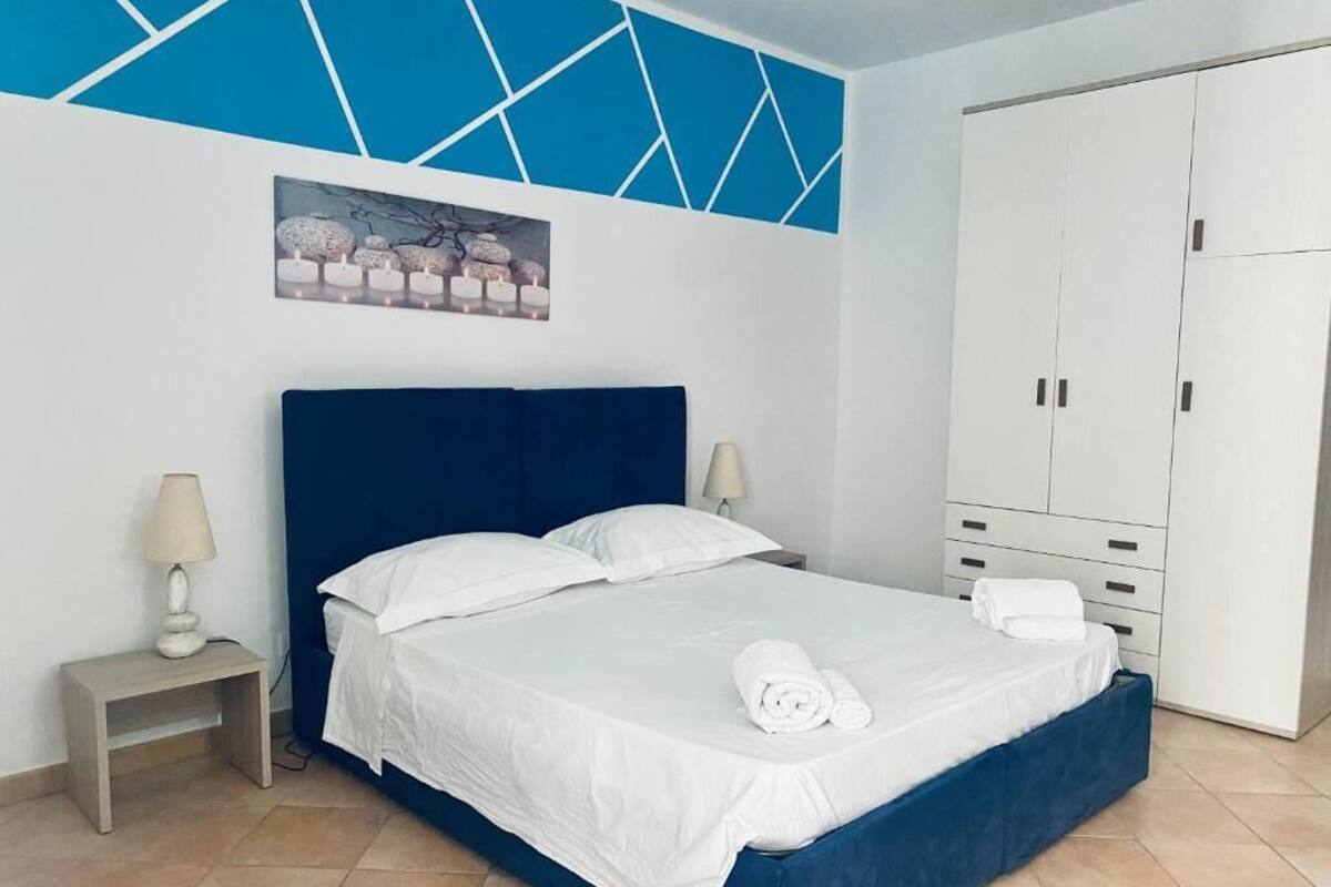 A neatly made bed with white linens and a rolled towel, a nightstand with a lamp, a painting on the wall, and a wardrobe with multiple doors.
