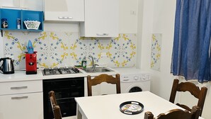 A compact kitchen with white cabinets, a gas stove, a sink, and a washing machine. There is a dining table with two chairs and a hanging light fixture. - CARTARI APARTMENT (Palermo)
