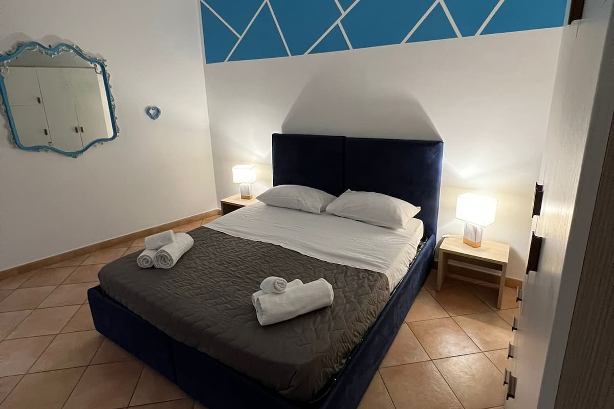 A neatly made bed with a dark headboard and white linens, a small bedside table with a lamp, and a geometric-patterned wall above the bed.
