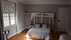 Un dormitorio con una cama grande, una mesita con plantas, una ventana con contraventanas y una puerta.