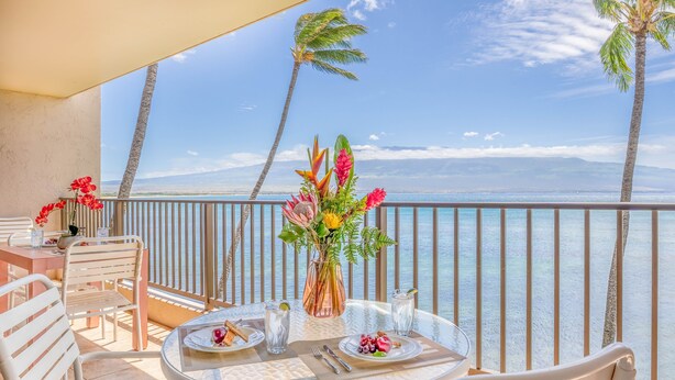 A balcony with a table set for two, featuring a vase with flowers, plates with food, and a view of the ocean and palm trees. - *Lauloa 312* Maui Prime 3 Bedroom Oceanfront Condo (Maalaea)