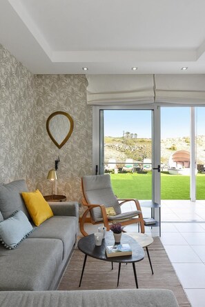 A modern living room with a grey sofa, a wooden armchair, and a coffee table. A large window offers a view of a green lawn and a stone wall.