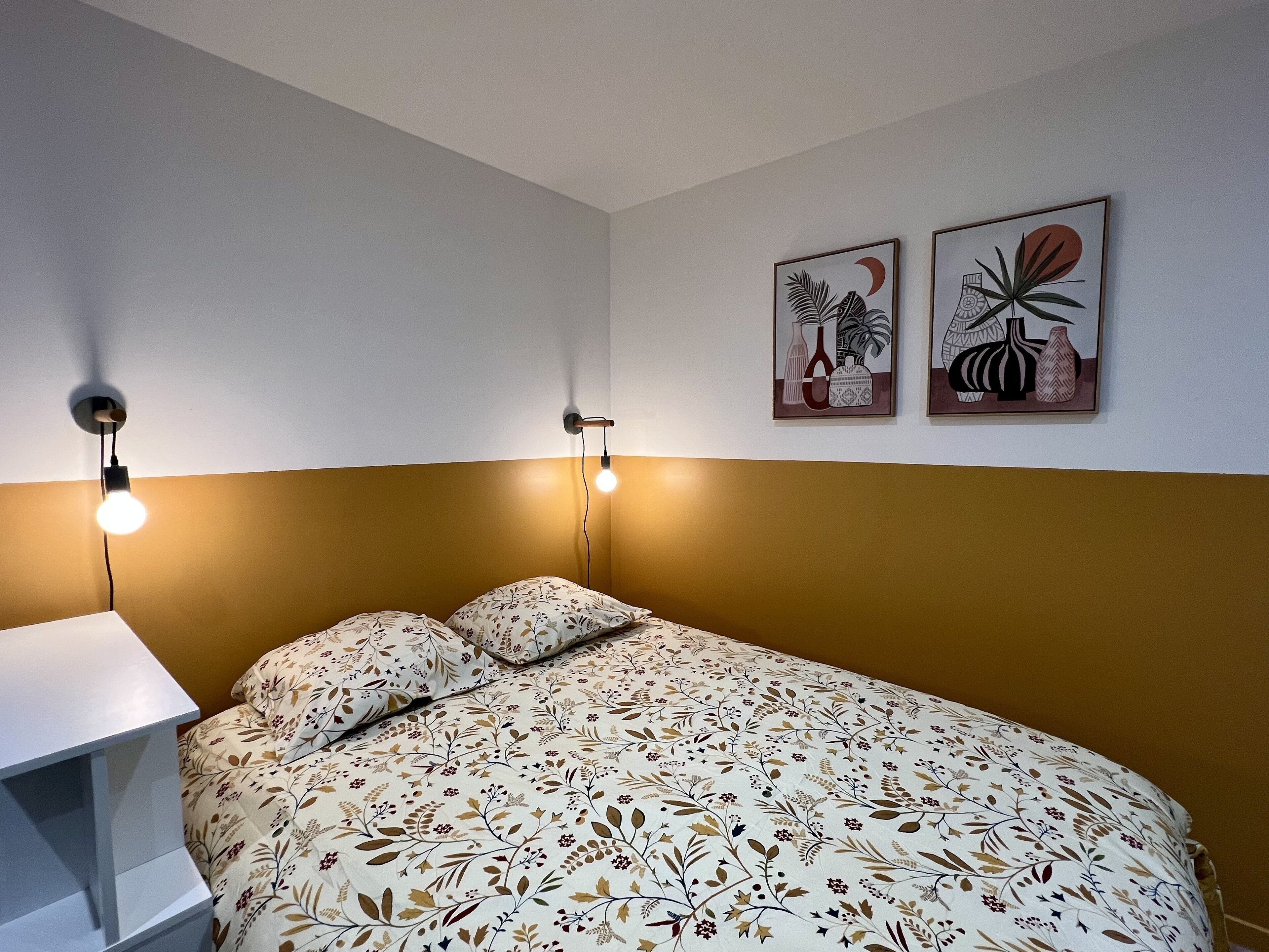 Une chambre à coucher avec un lit, deux lampes fixées au mur et deux œuvres d’art encadrées accrochées au mur.