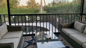 Et overdækket udendørs område med fletningsmøbler, et bord med glasplade og udsigt til pool og grøn bevoksning.