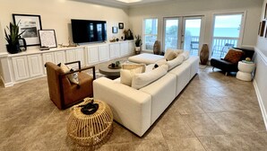 A spacious living room with a large white sofa, a brown armchair, and a round ottoman. A flat-screen TV is mounted on the wall, and there is a ceiling fan above. - Solaria: Waterfront, Heated Pool, Kayak, Fish/Golf, St George Island (Eastpoint)