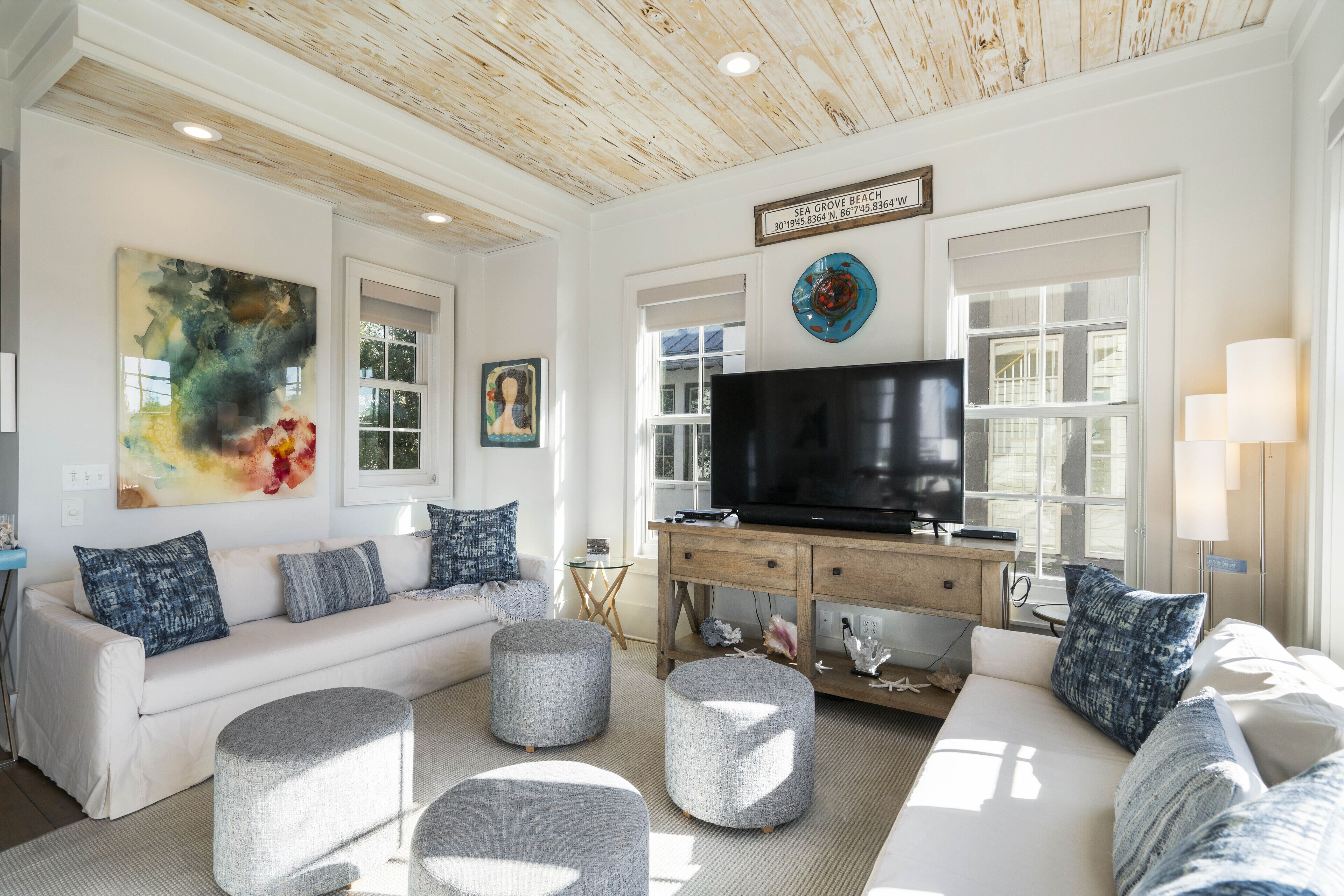 A modern living room with a white sofa, grey ottomans, a flat-screen TV, and a wooden ceiling.