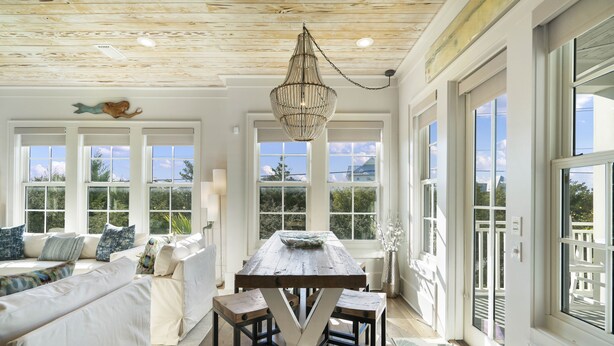 Una habitación amplia con techo de madera, ventanas grandes, una mesa de comedor con taburetes y una zona de estar acogedora con cojines.