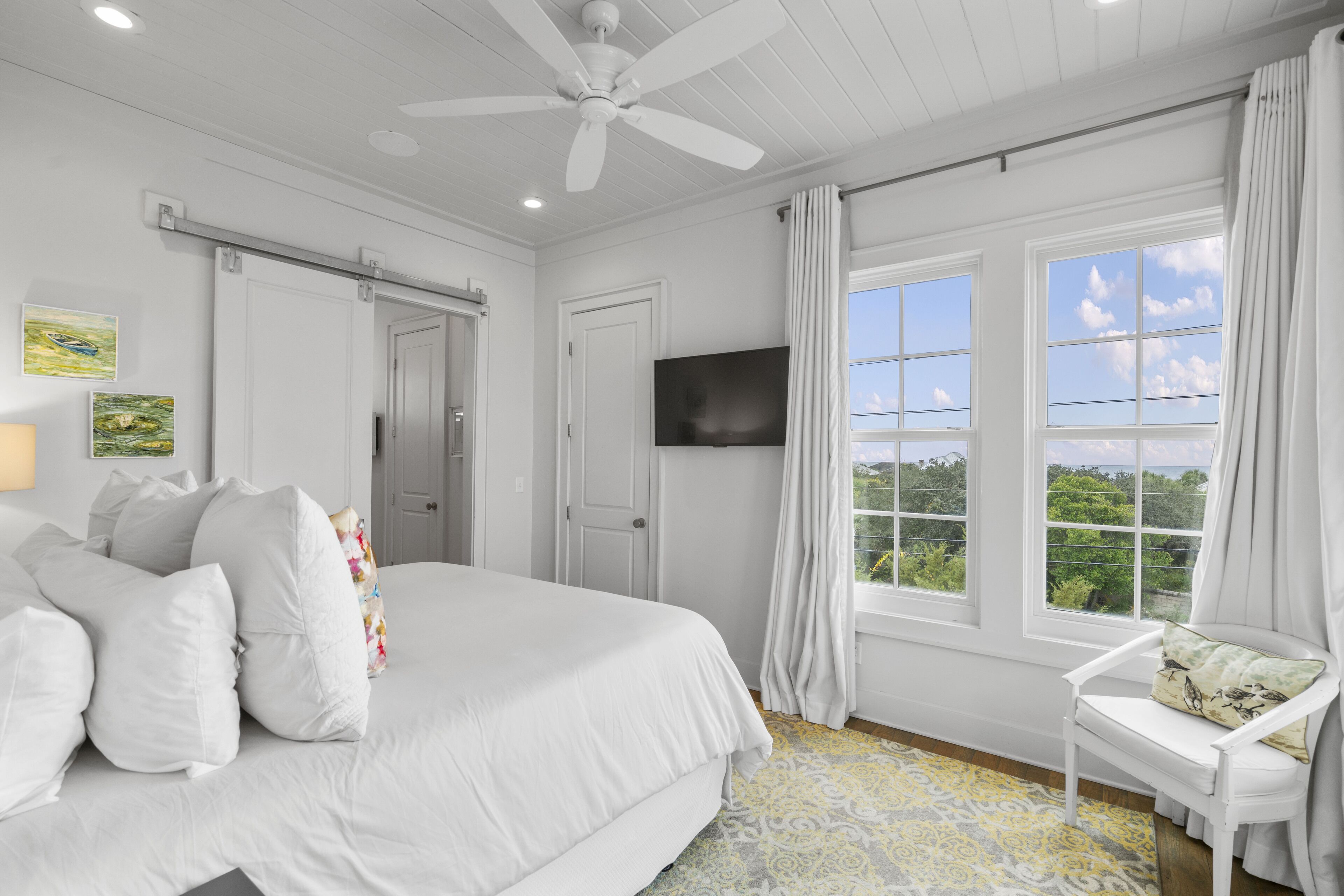 A bedroom with a bed, a chair, a ceiling fan, a TV, and a window with curtains.