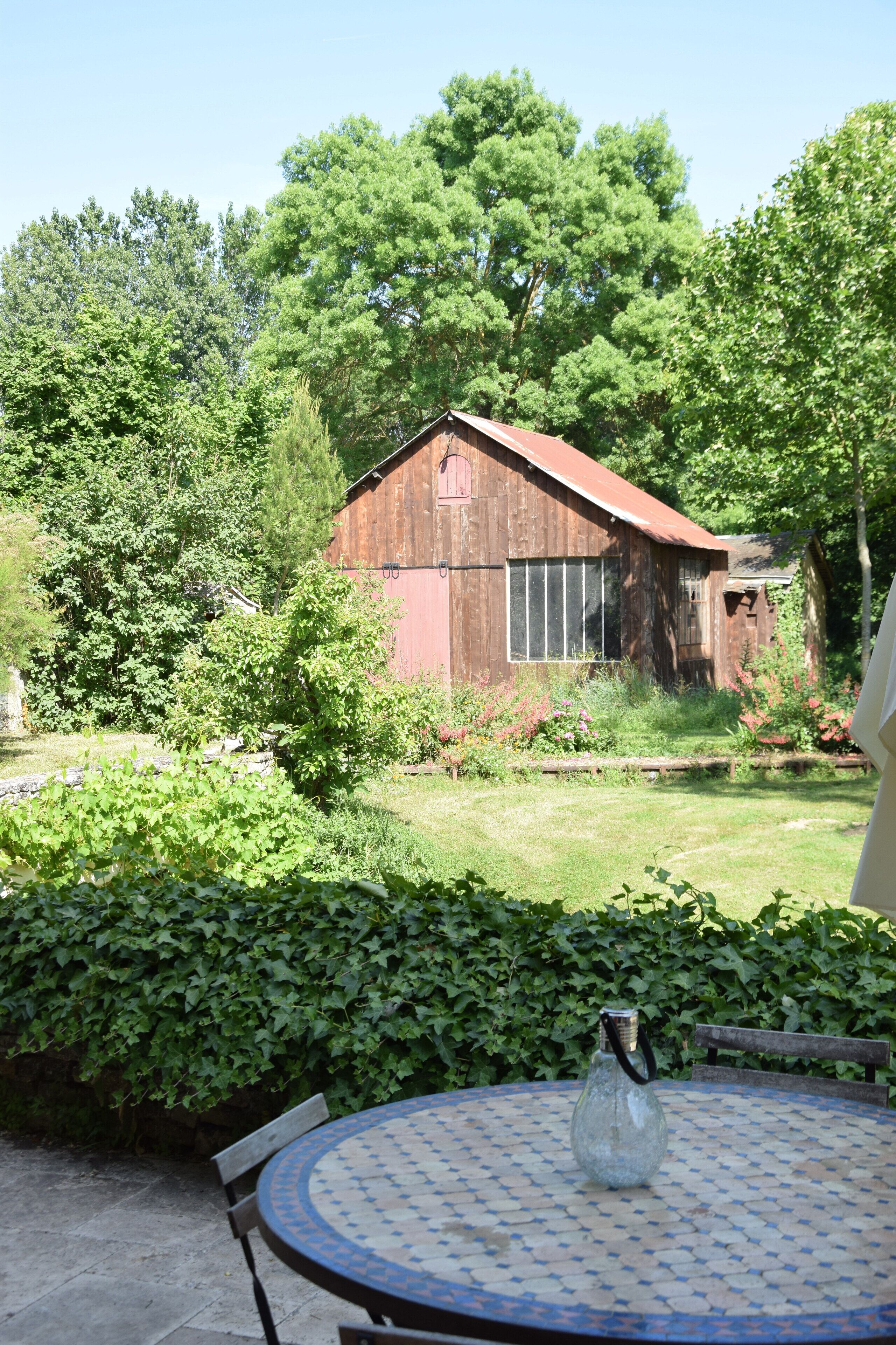 Une grange rustique en bois, avec un toit rouge, entourée d’une végétation luxuriante et d’un jardin bien entretenu.