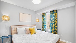 Un dormitorio con una cama grande, dos mesitas de noche, una lámpara de pared, una ventana con cortinas e imagen enmarcada en la pared.