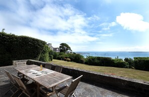 An outdoor patio with a wooden table and chairs, a stone wall, and a view of the sea and a distant building.